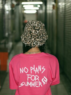 Back view of unrecognizable female teenager in stylish hat and t shirt with No Plans For Summer inscription standing in corridor of modern building