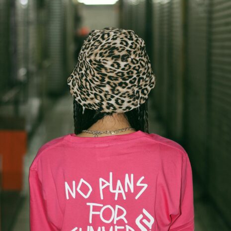 Back view of unrecognizable female teenager in stylish hat and t shirt with No Plans For Summer inscription standing in corridor of modern building